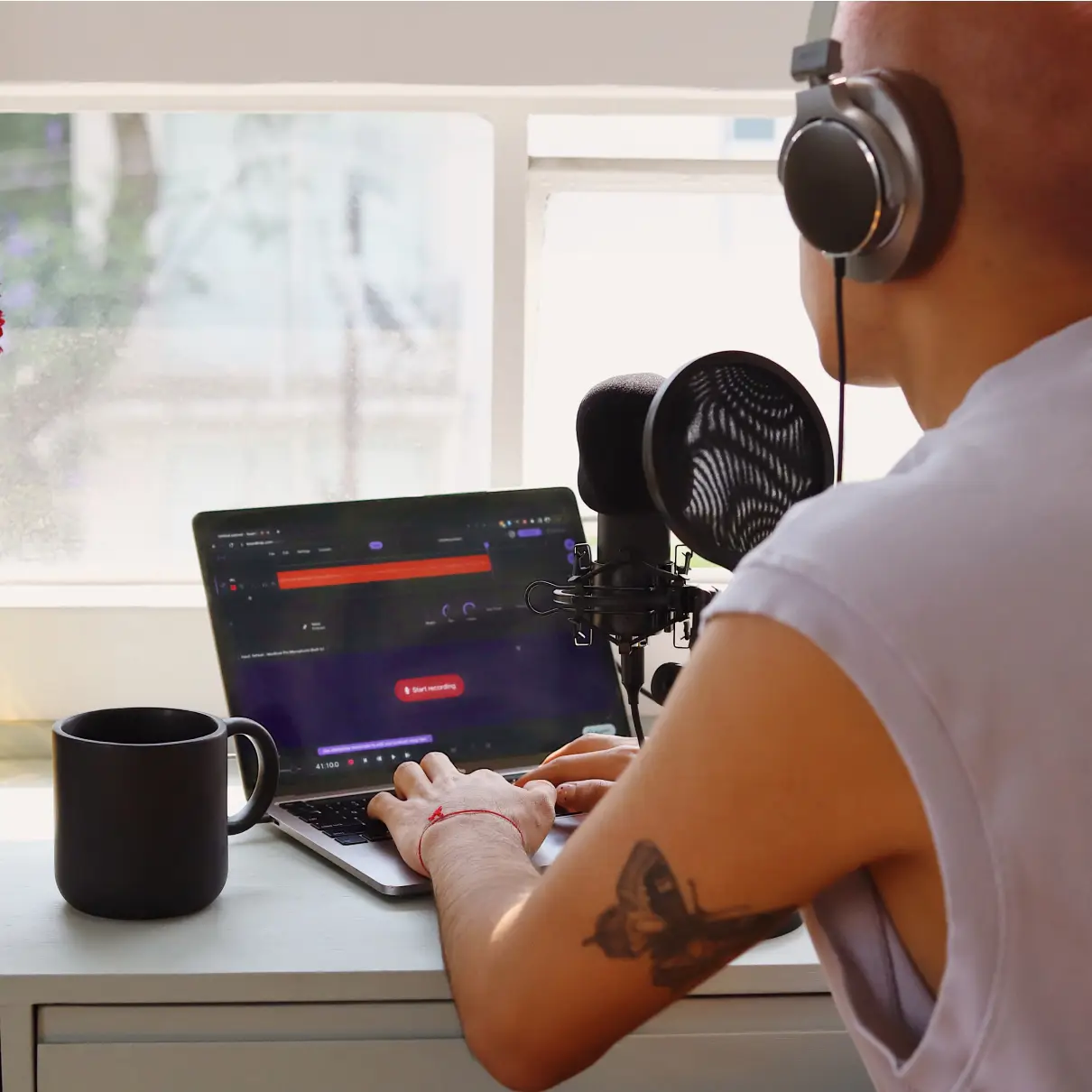 The back of someone who is looking at a screen with the Soundtrap Studio in dark mode on. They have a coffee cup beside them, and a microphone in front of them. The person is wearing headphones.