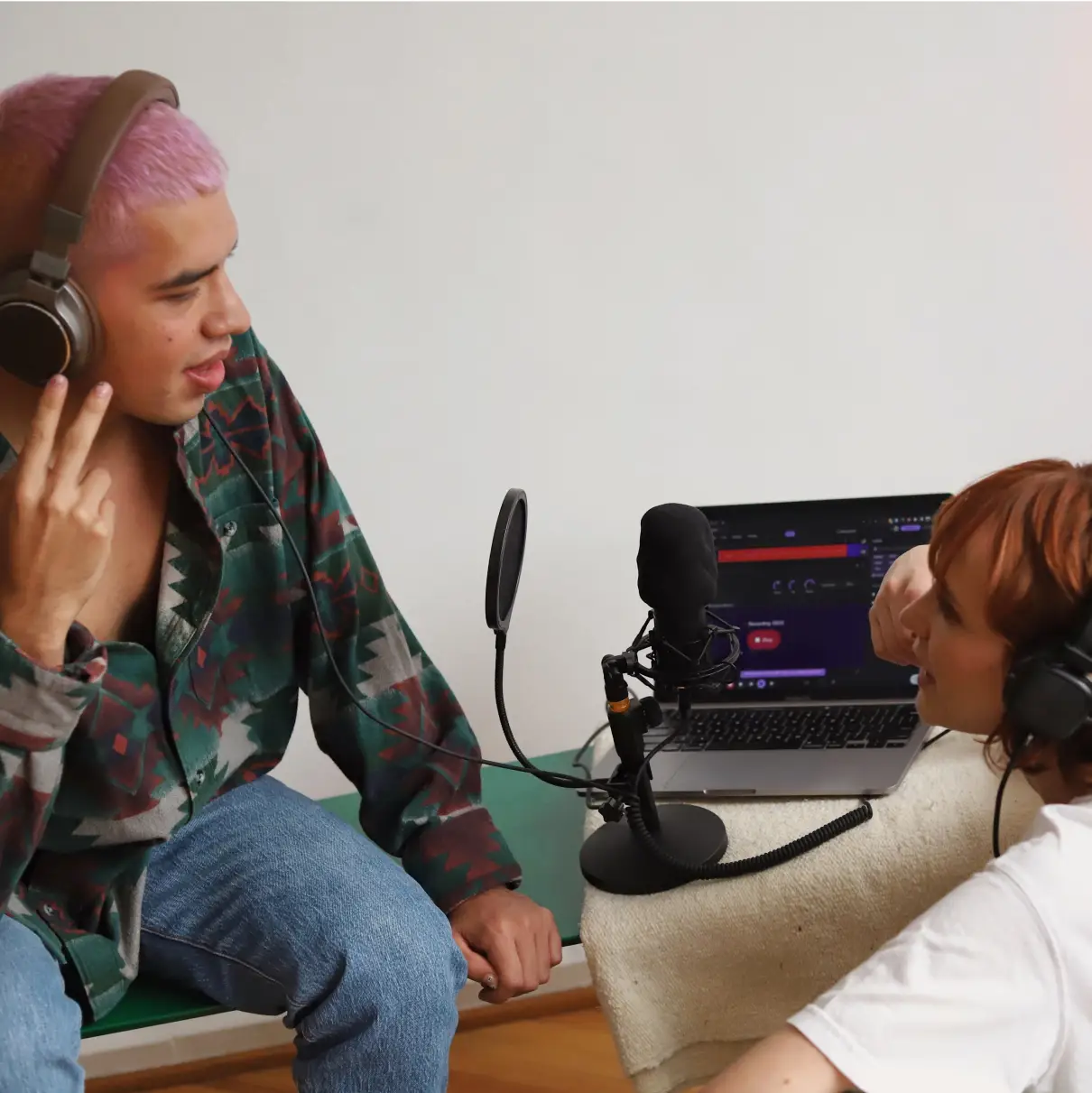 Two people are looking at each other with a microphone placed between them. They are recording a podcast. They are both wearing headphones. The Soundtrap Studio is open in dark mode on a laptop behind them.