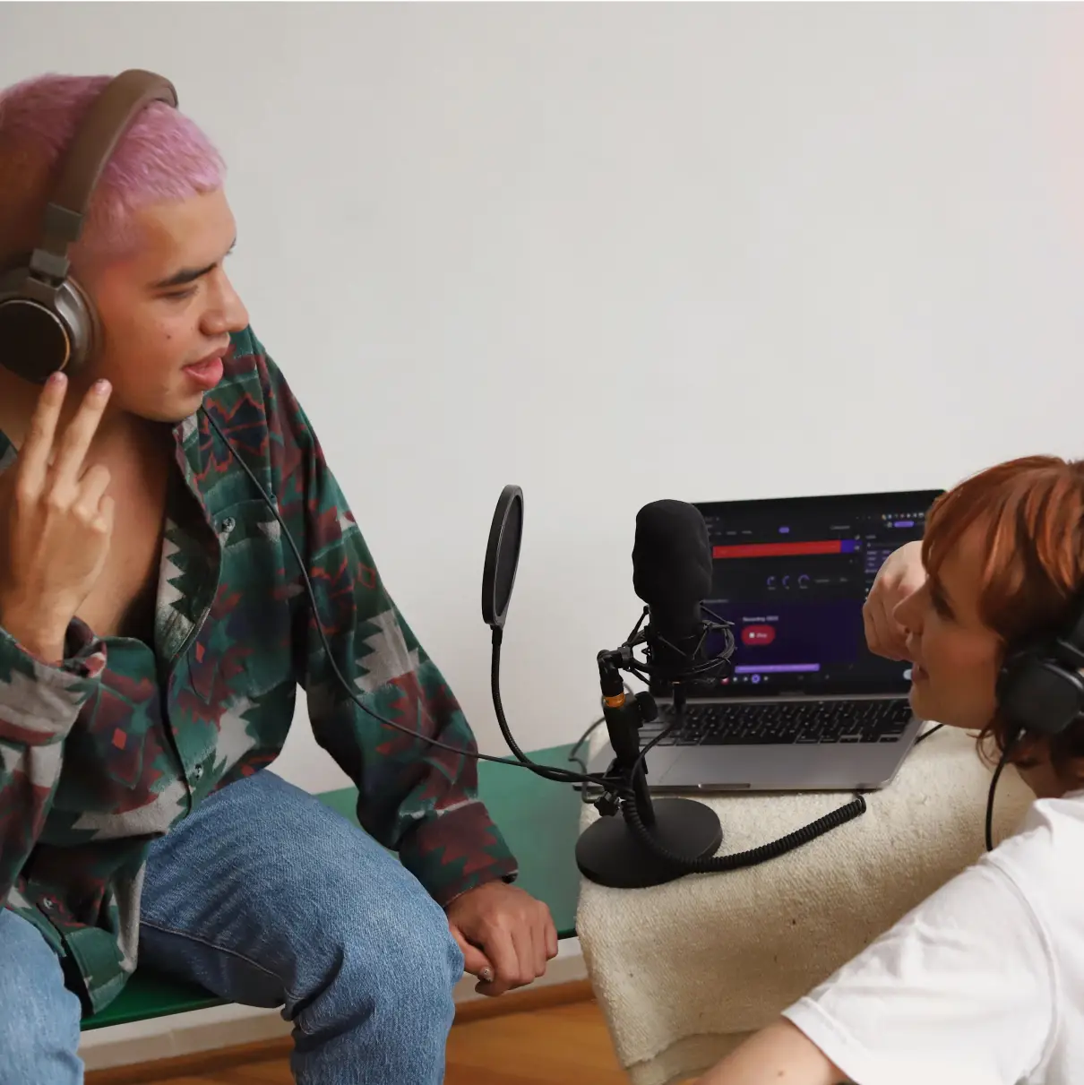 Dua orang saling memandang dengan mikrofon di antara mereka. Mereka sedang merekam podcast. Mereka berdua memakai headphone. Soundtrap Studio terbuka dalam mode gelap di laptop di belakang mereka.